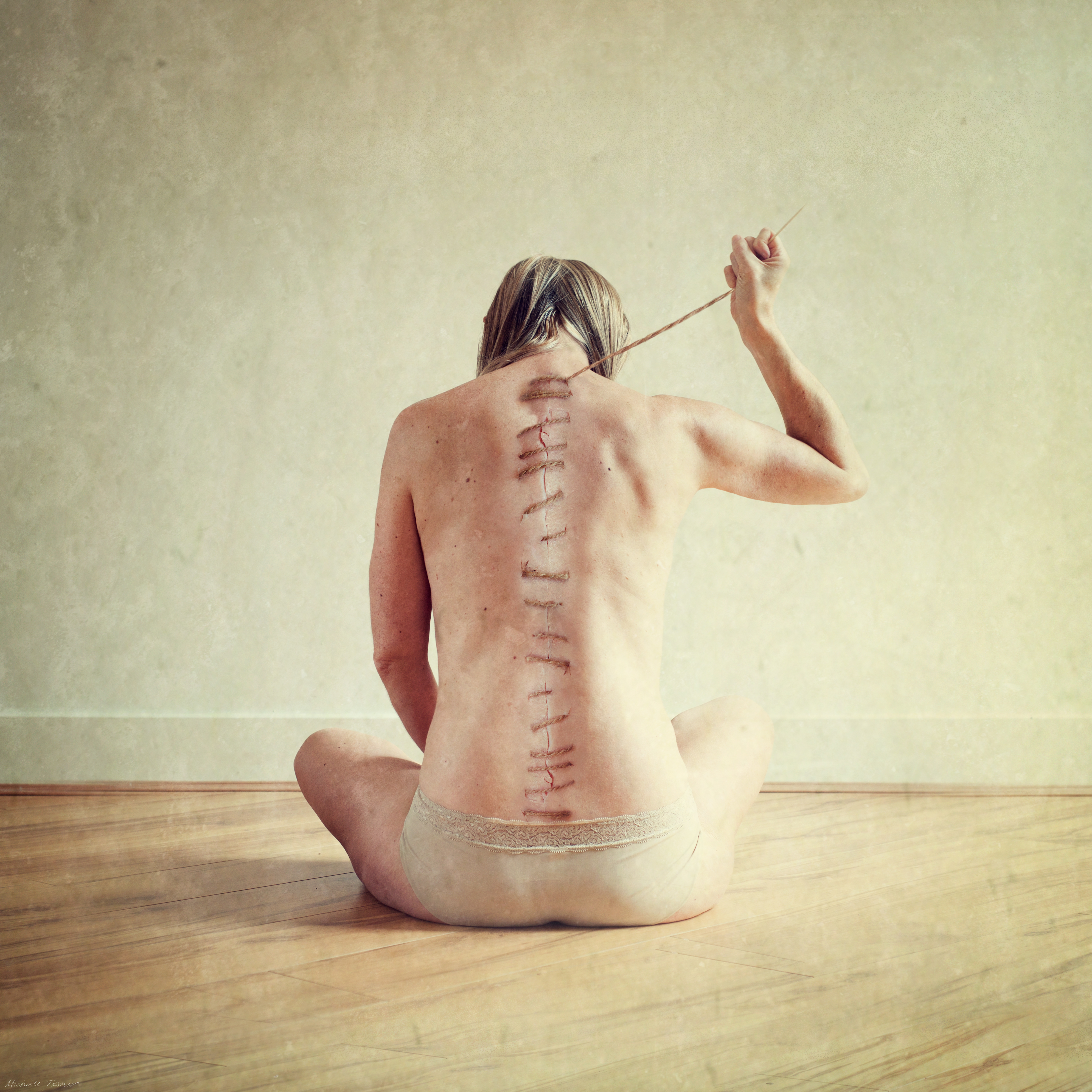 A woman sits facing a wall, her naked back to the camera. Coarse, brown twine stitches run up her spine. At the top of her spine she holds the needle and thread in her right hand.
